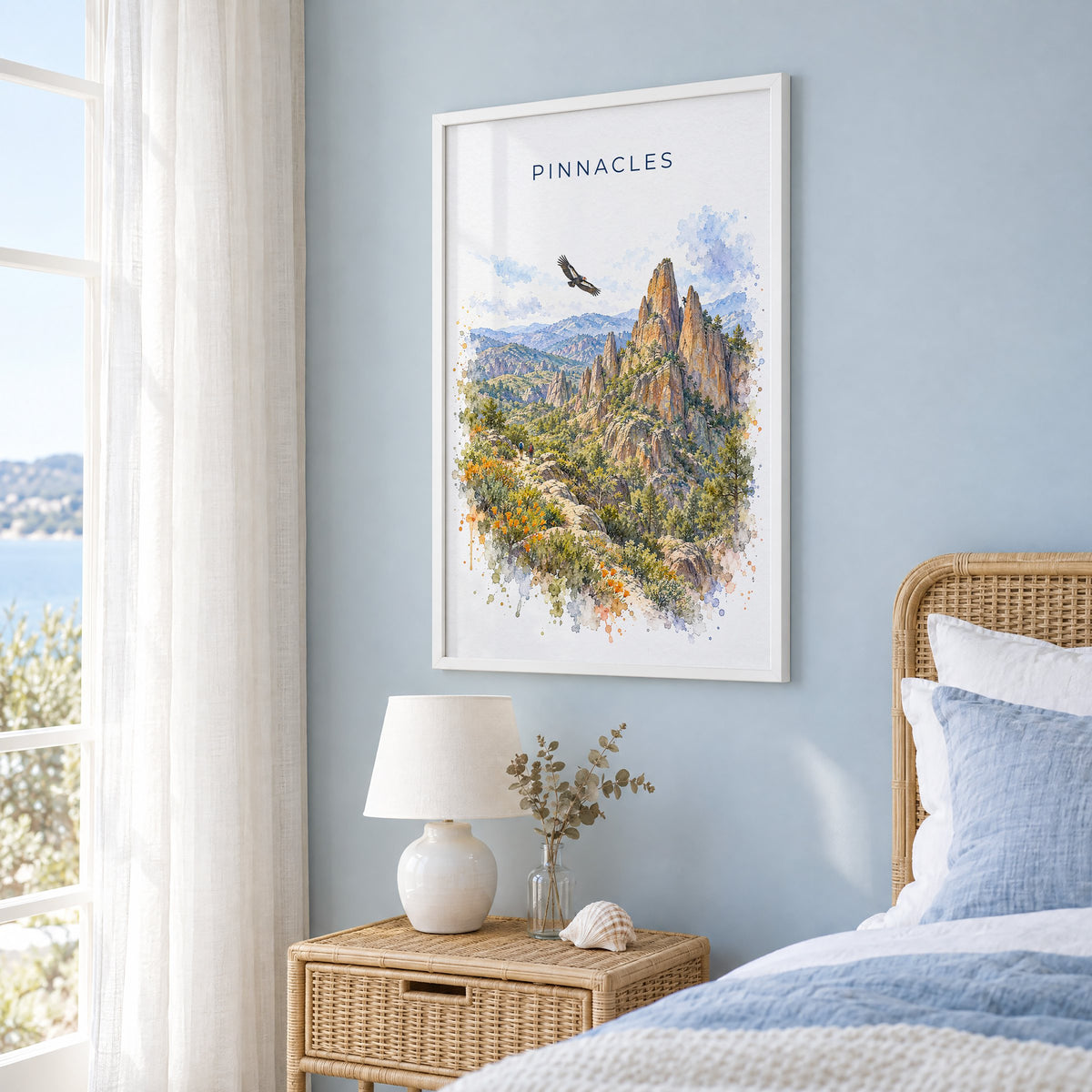 Pinnacles National Park watercolor print in a white frame on a pale blue wall beside a rattan bed in a coastal bedroom