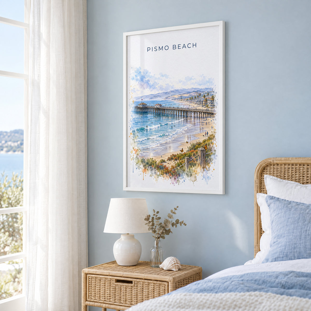 Pismo Beach watercolor print in a white frame on a pale blue wall beside a rattan bed in a coastal bedroom