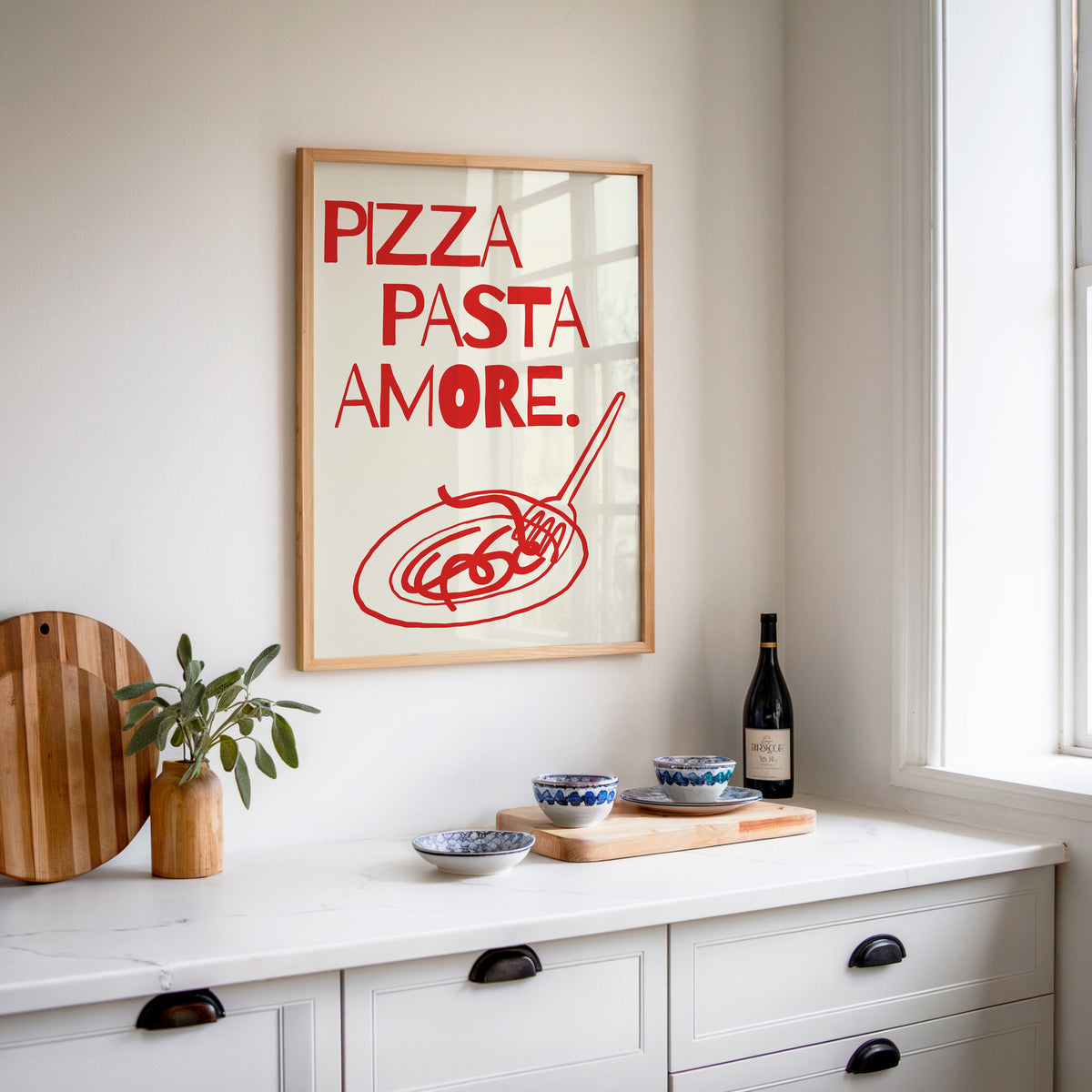 Pizza Pasta Amore print in an oak frame above a white sideboard with a bottle of wine