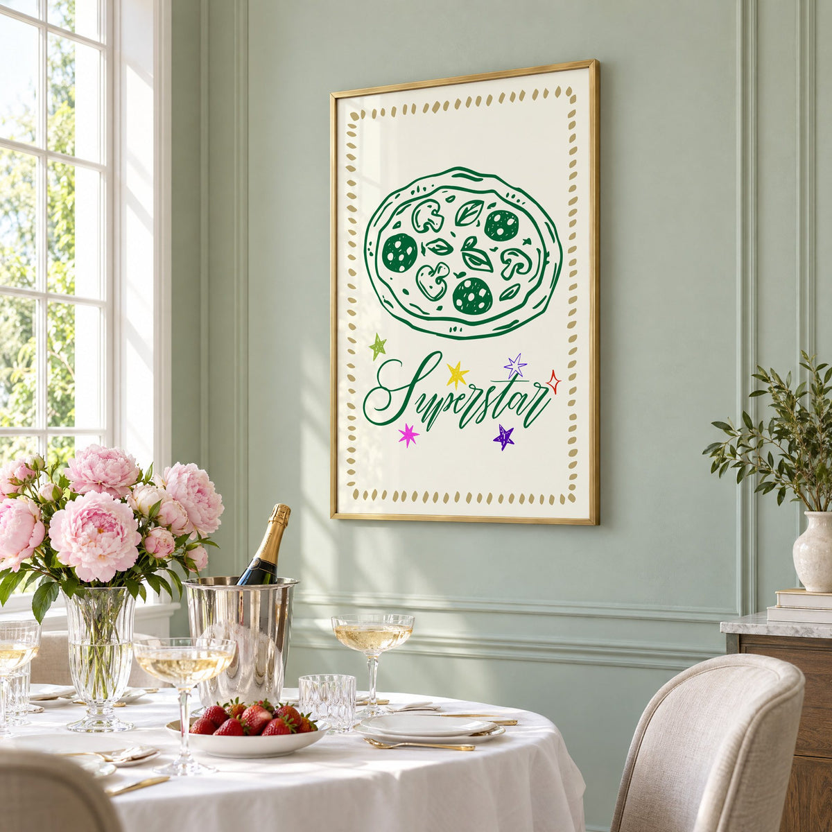 Superstar green pizza and stars print in a brass frame on a sage wall above a brunch table with champagne and peonies, seen at an angle