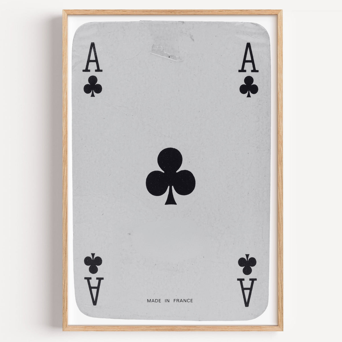 Worn vintage ace of clubs playing card print in a light oak frame on a plain white wall