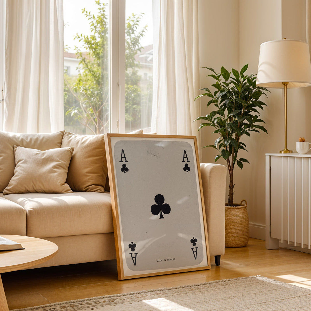 Worn vintage ace of clubs playing card print in a light oak frame leaning on a cream sofa beside a floor lamp
