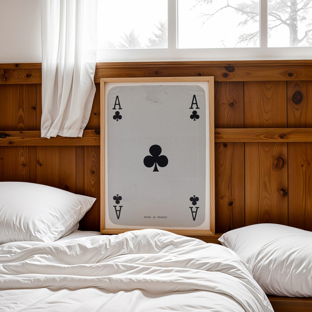Worn vintage ace of clubs playing card print in a pine frame leaning on a wood headboard over white bedding