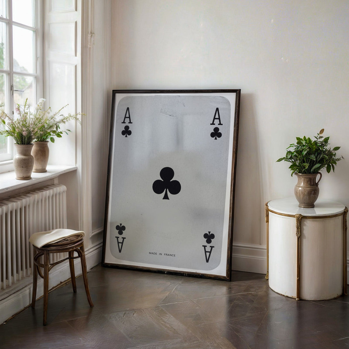 Worn vintage ace of clubs playing card print in a black frame on a dark stone floor beside a white side table