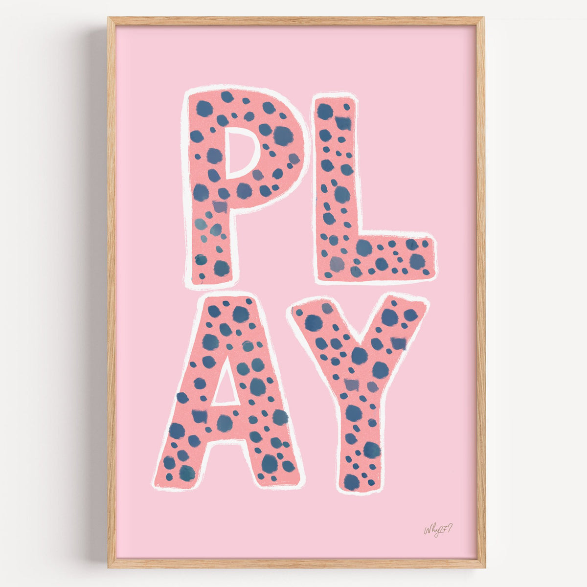 Spotted play letters pink nursery print in a light oak frame on a plain white wall