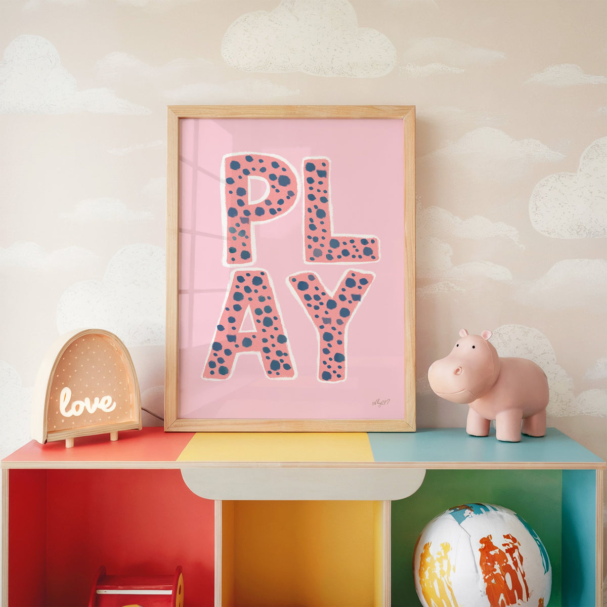 Spotted play letters pink nursery print in an oak frame on a rainbow toy shelf against cloud wallpaper