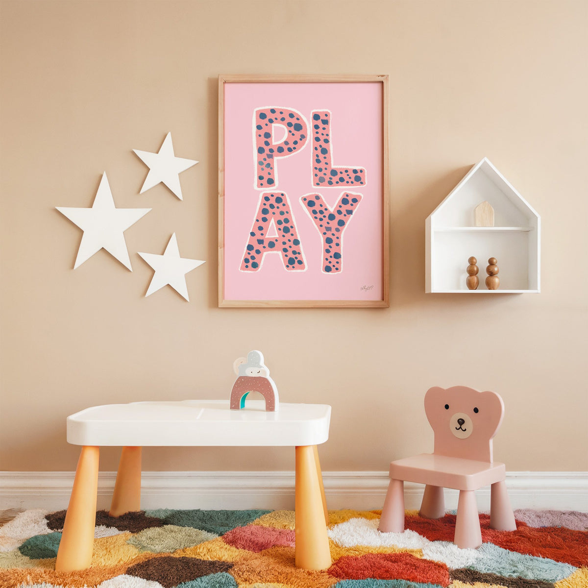 Spotted play letters pink nursery print in an oak frame on a peach wall above a kids table with wall stars