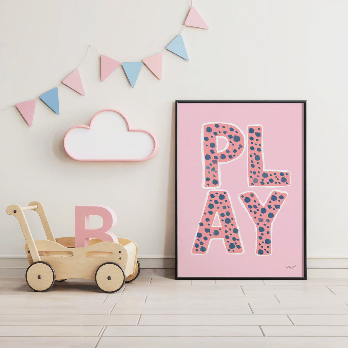 Spotted play letters pink nursery print in a black frame under pastel bunting by a wooden baby walker
