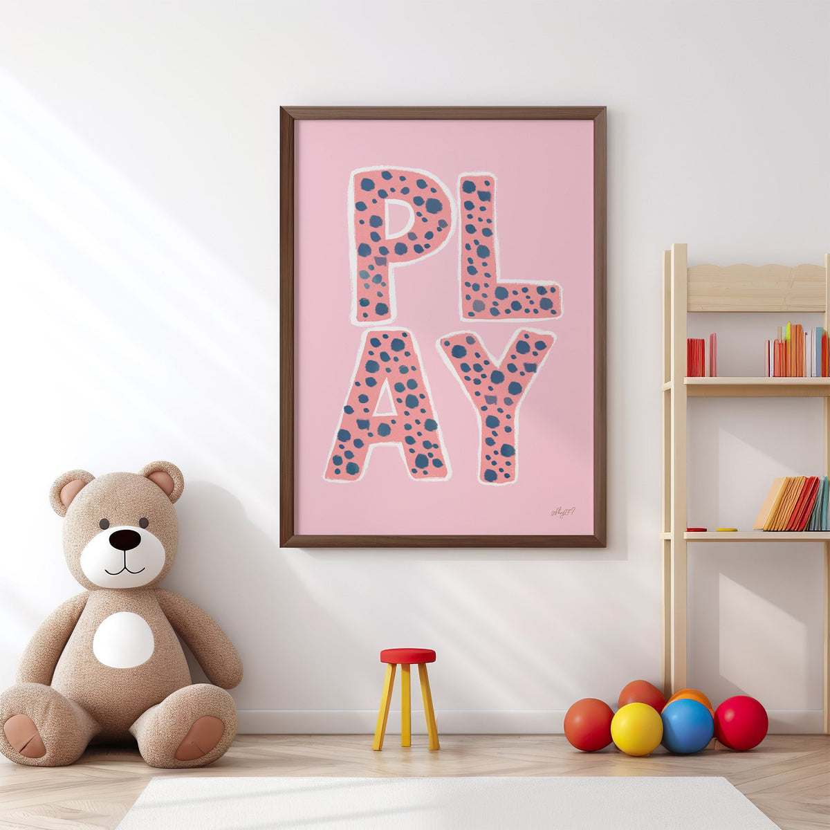 Spotted play letters pink nursery print in a walnut frame above a big teddy bear by a bookshelf
