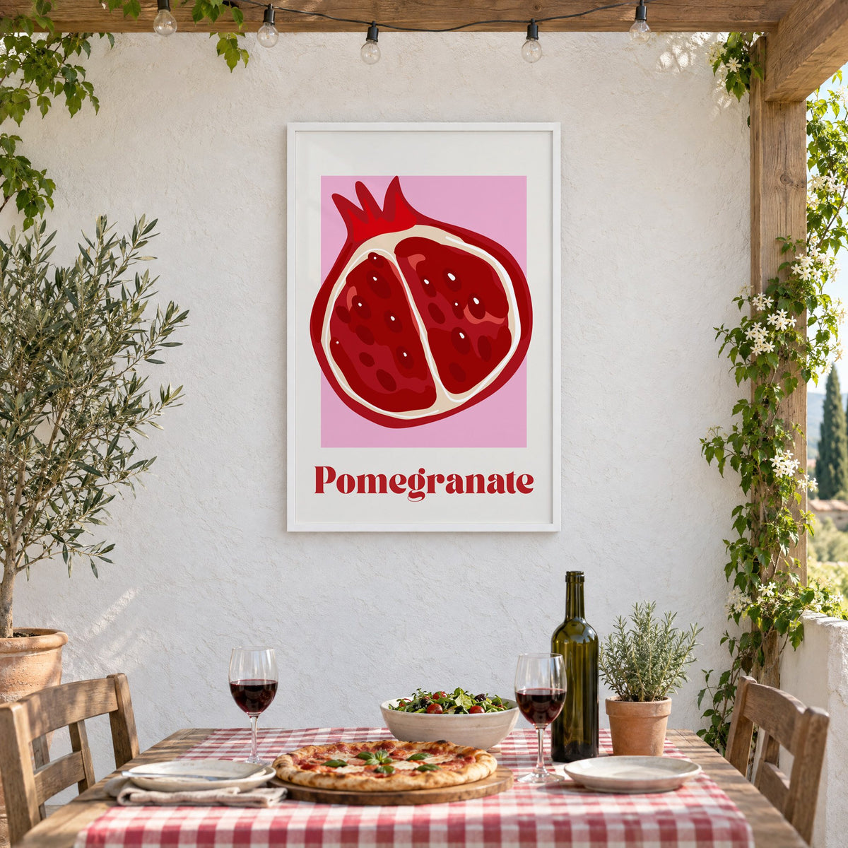 Pomegranate half bold serif pink panel print in a white frame on a stucco wall above a checked-cloth table with pizza and red wine