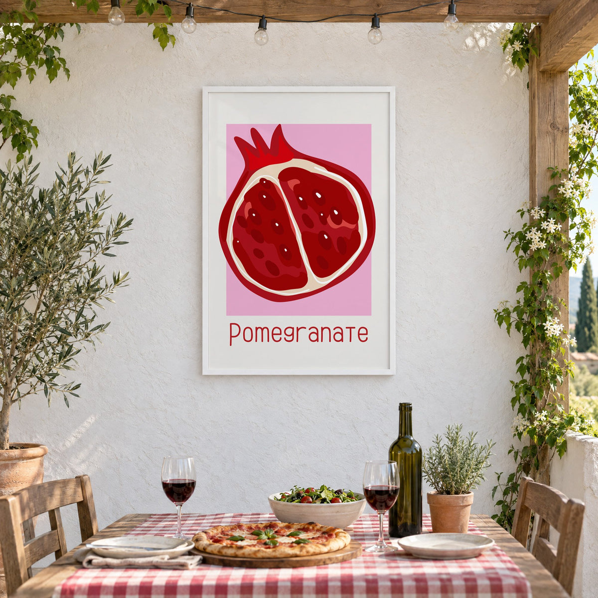 Pomegranate thin lettering halved fruit print in a white frame on a stucco wall above a checked-cloth table with pizza and red wine
