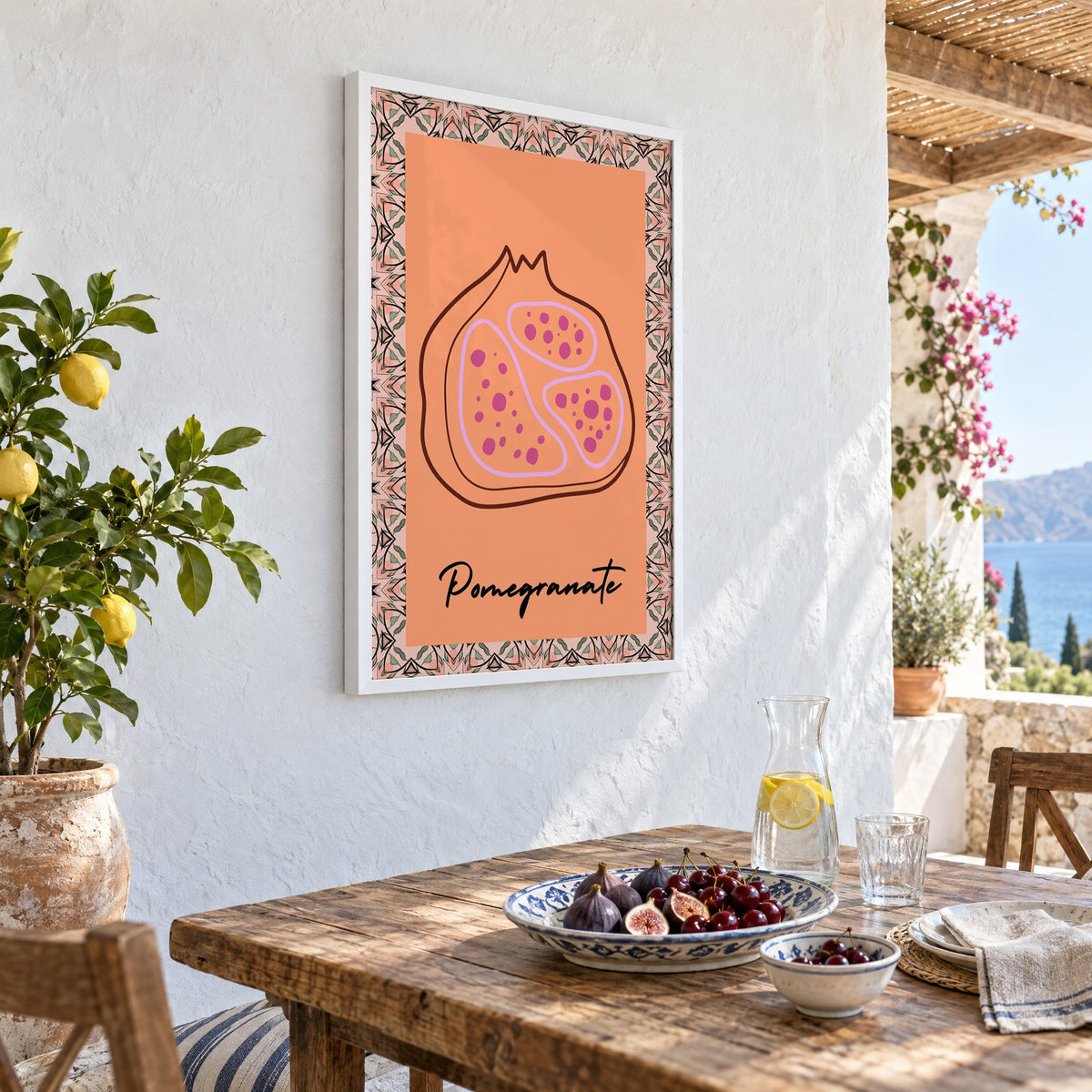 Pomegranate line-art kaleidoscope border print in a white frame on a whitewashed terrace wall above a lunch table with figs and cherries, seen at an angle