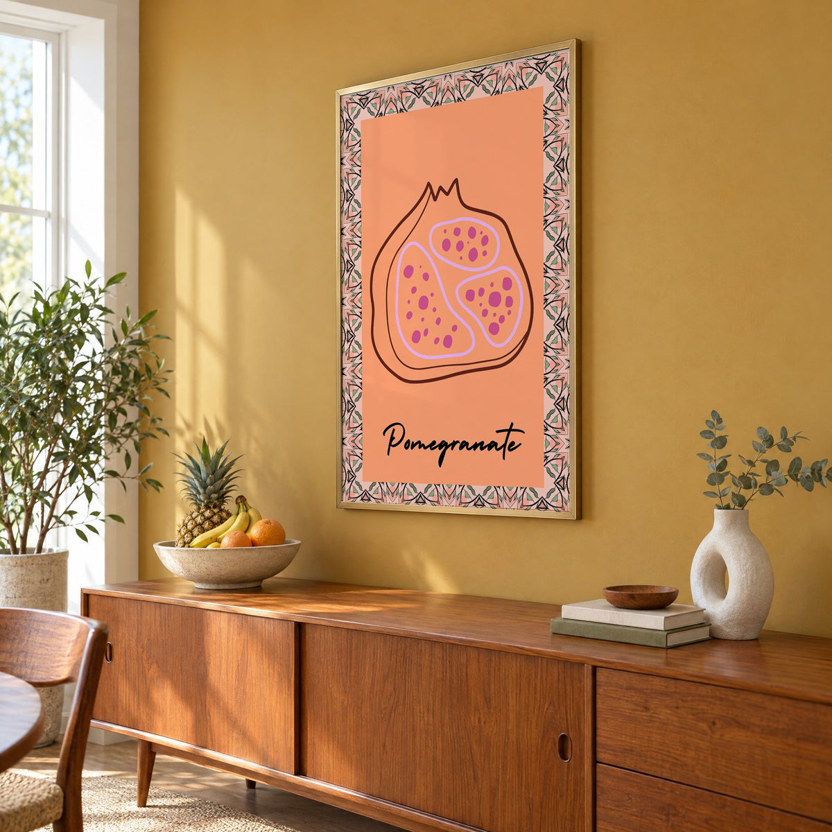 Pomegranate line-art kaleidoscope border print in a brass frame on a mustard wall above a teak sideboard with a fruit bowl, seen at an angle