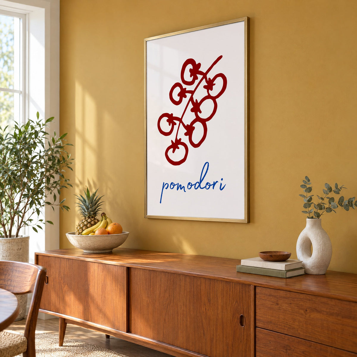 pomodori red cherry tomato vine print in a brass frame on a mustard wall above a teak sideboard with a fruit bowl, seen at an angle