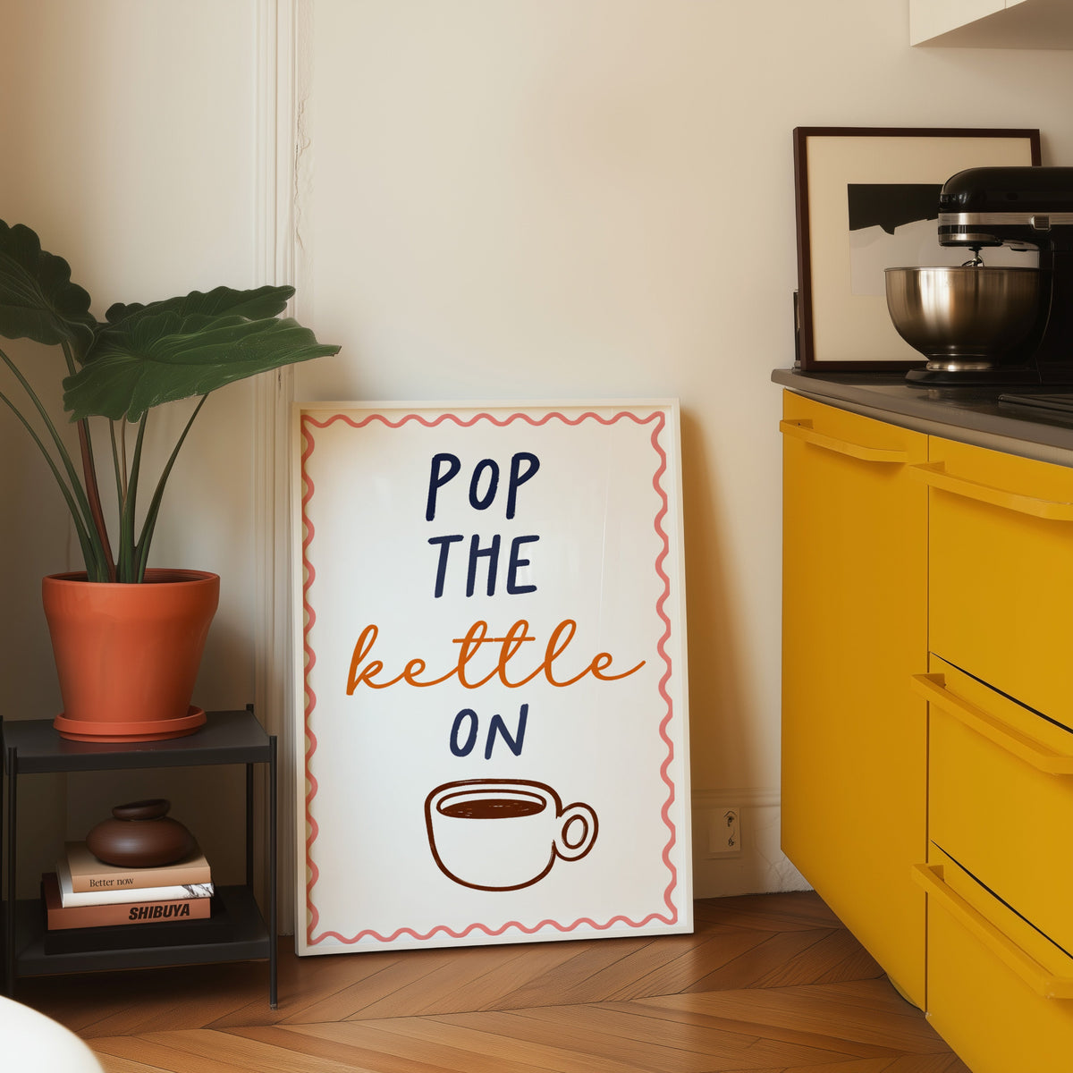 Pop the Kettle On print in a white frame on parquet beside a bright yellow kitchen