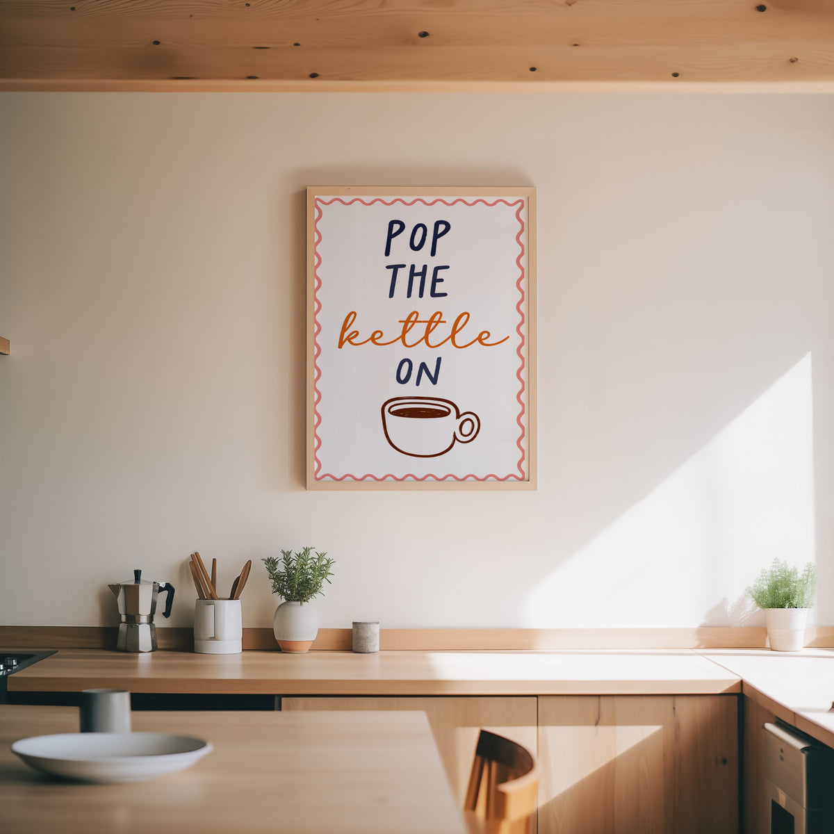 Pop the Kettle On print in an oak frame above a wood counter with a moka pot