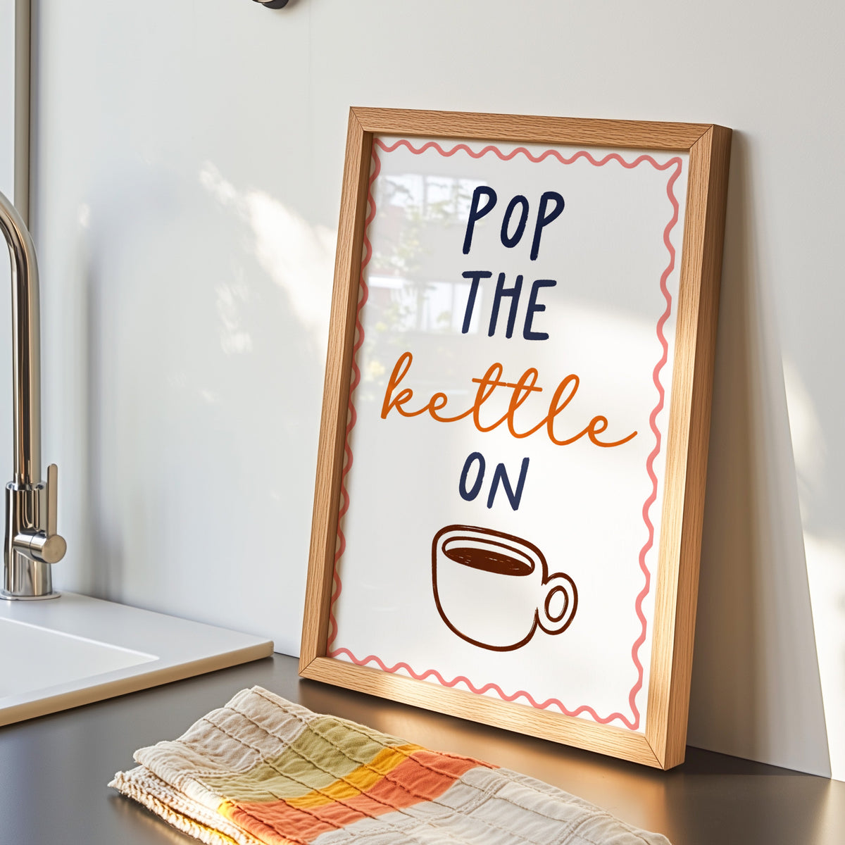 Pop the Kettle On print in an oak frame by the sink with a striped tea towel
