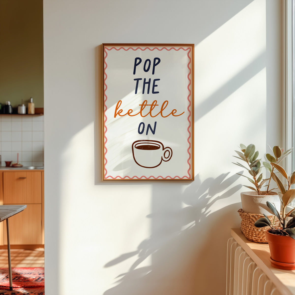 Pop the Kettle On print in a gold frame in sunlight with plants and a kitchen beyond