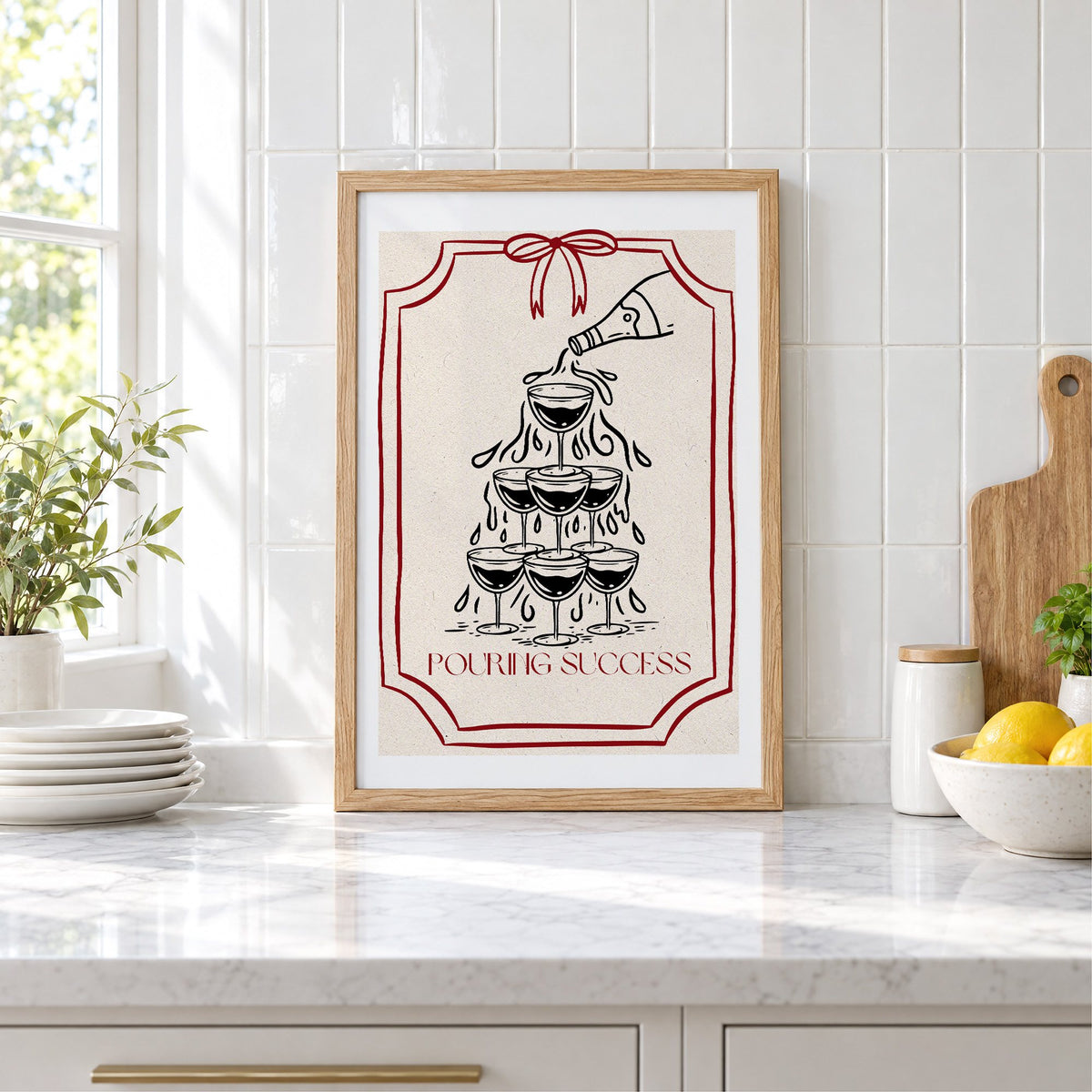 POURING SUCCESS champagne tower print in an oak frame leaning on a marble counter by white tiles and lemons