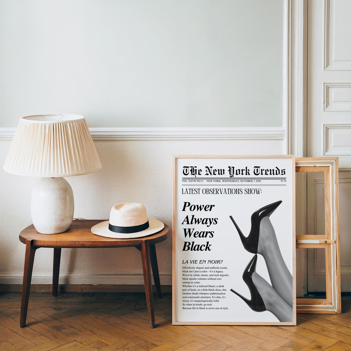 Two pairs of black heels mock newspaper print in a pine frame on a wood floor beside a lamp and straw hat