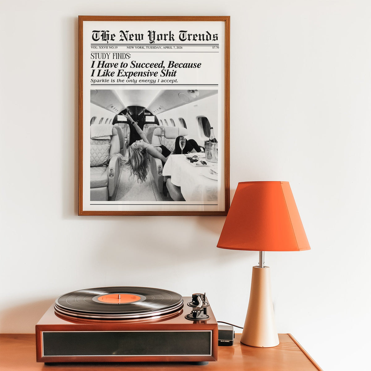 Woman with champagne in private jet mock newspaper print in a walnut frame above a record player and orange lamp