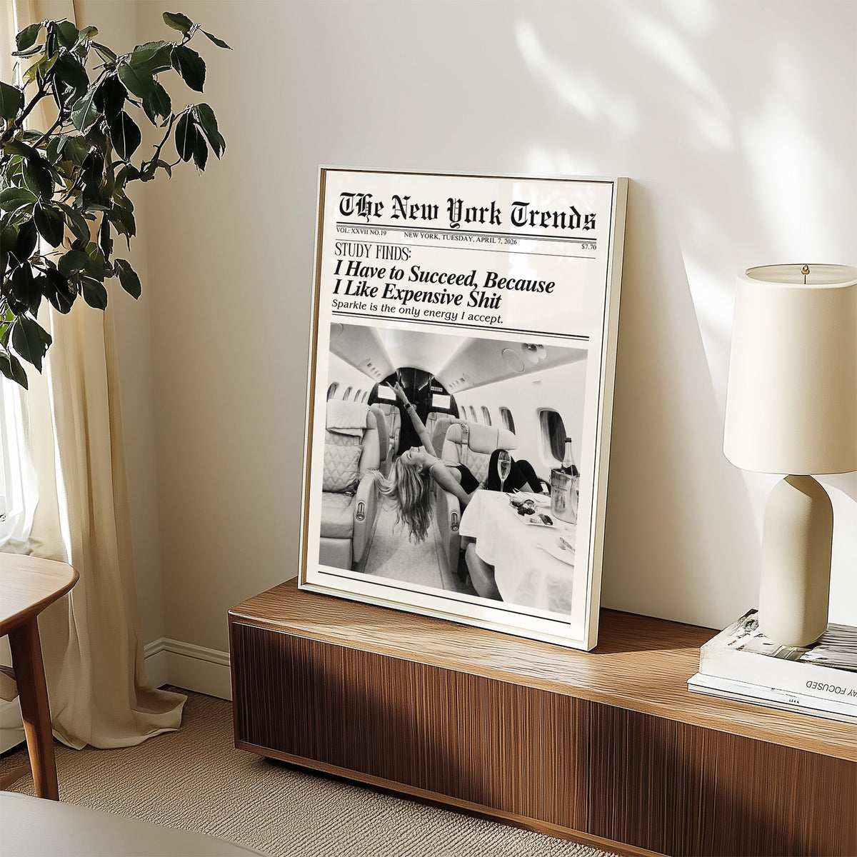 Woman with champagne in private jet mock newspaper print in a white frame on a walnut sideboard beside a ceramic lamp