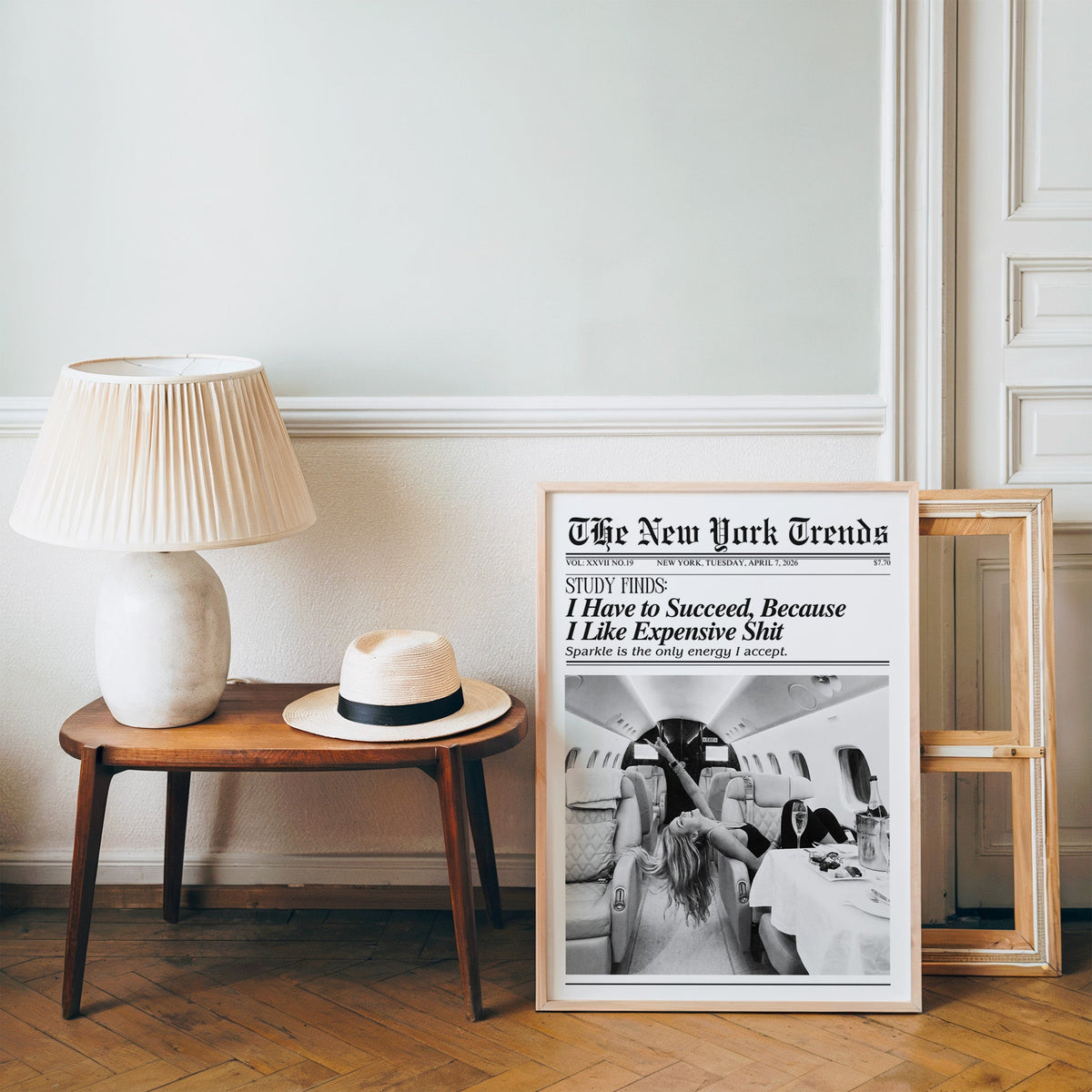 Woman with champagne in private jet mock newspaper print in a pine frame on a wood floor beside a lamp and straw hat