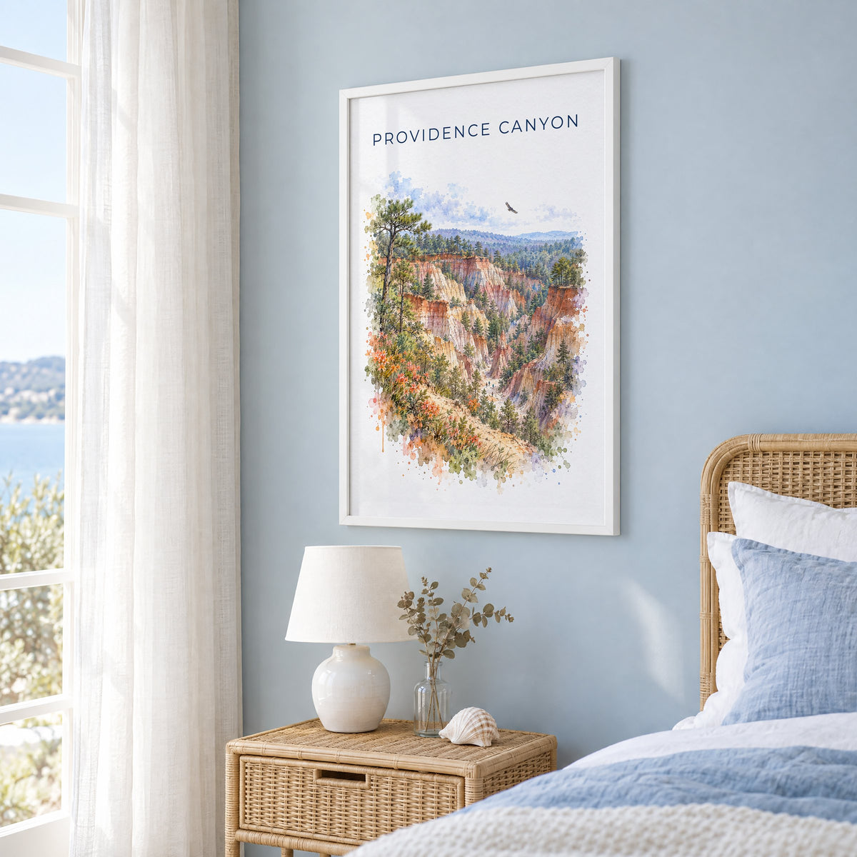 Providence Canyon State Park watercolor print in a white frame on a pale blue wall beside a rattan bed in a coastal bedroom