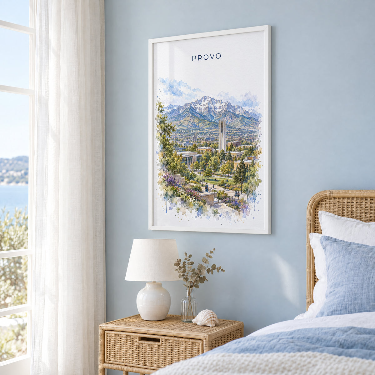 Provo watercolor print in a white frame on a pale blue wall beside a rattan bed in a coastal bedroom
