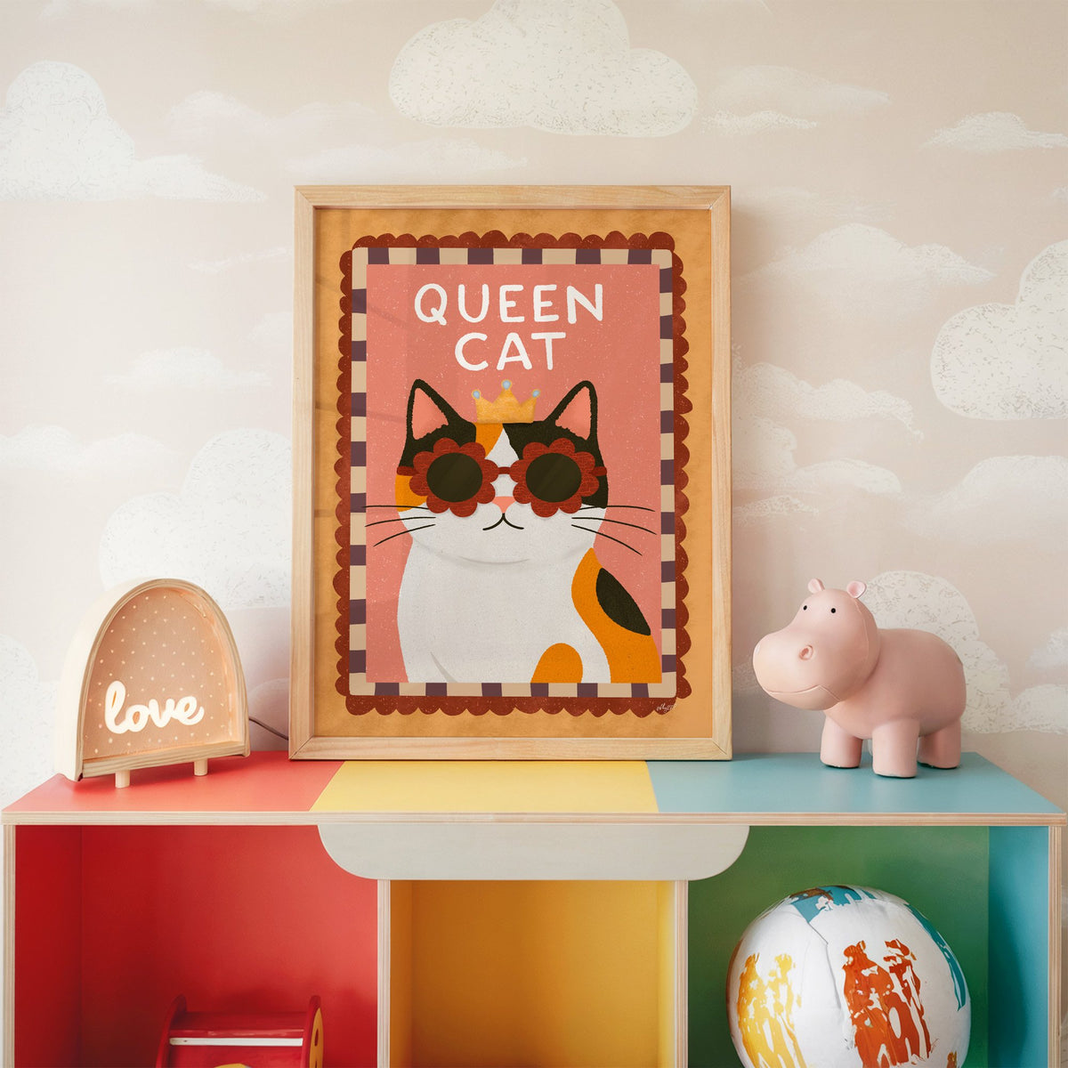 Calico cat with gold crown nursery print in an oak frame on a rainbow toy shelf against cloud wallpaper
