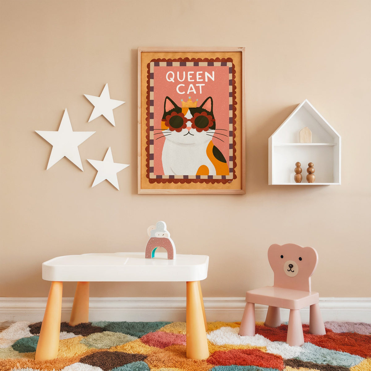 Calico cat with gold crown nursery print in an oak frame on a peach wall above a kids table with wall stars