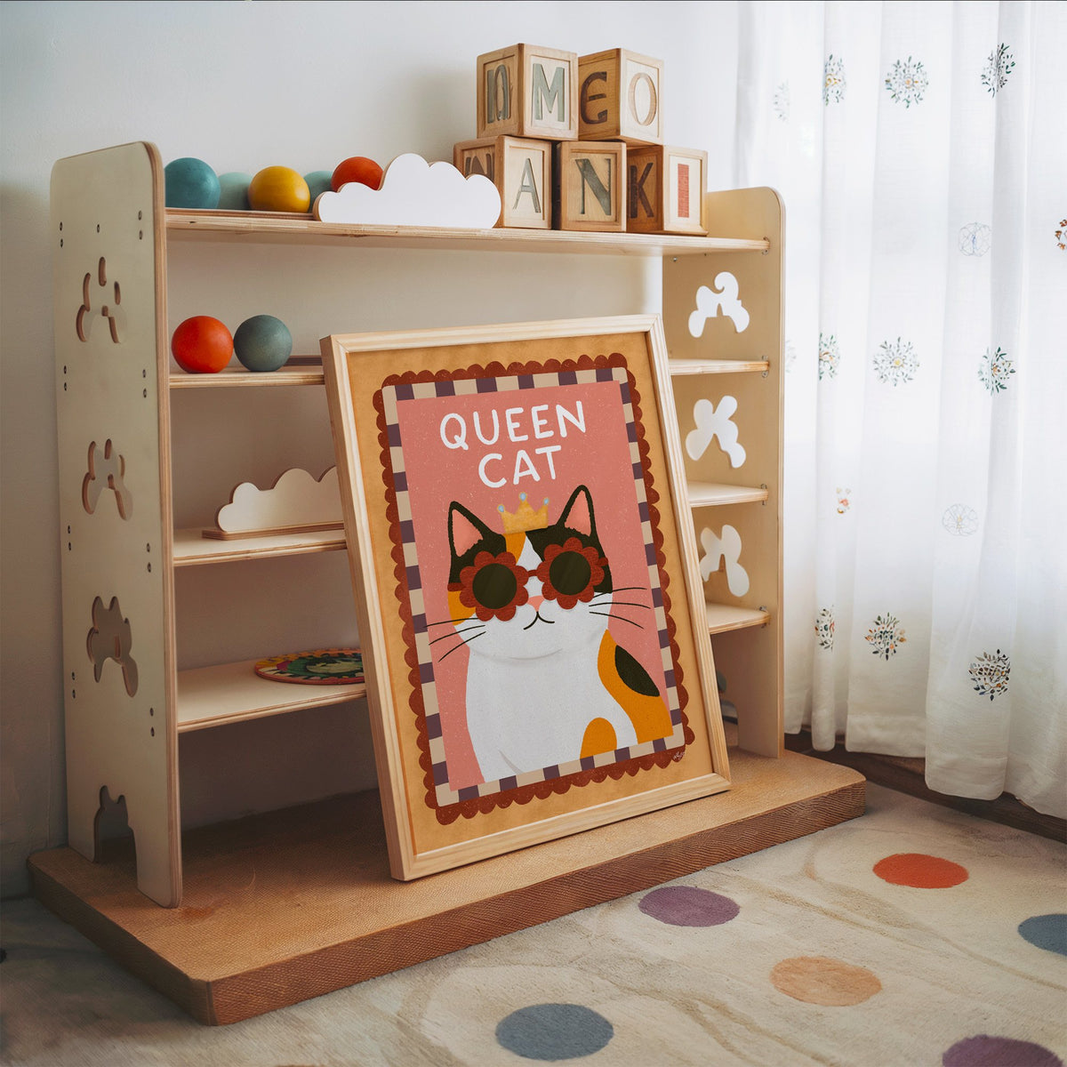 Calico cat with gold crown nursery print in an oak frame leaning on a toy shelf with wooden letter blocks