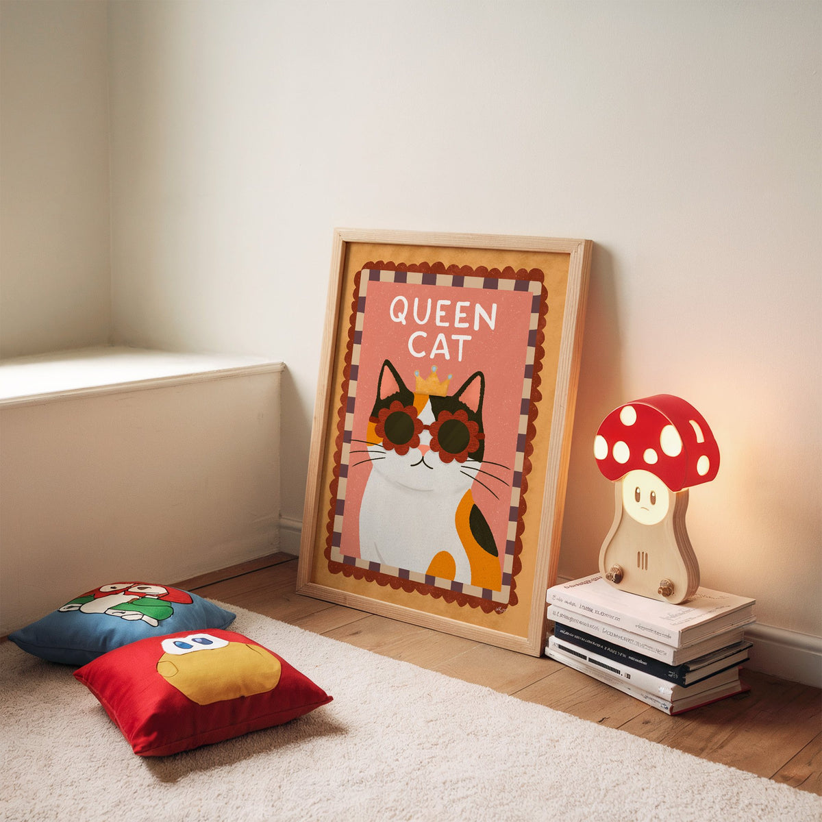 Calico cat with gold crown nursery print in an oak frame leaning on a rug beside a mushroom lamp and floor cushions