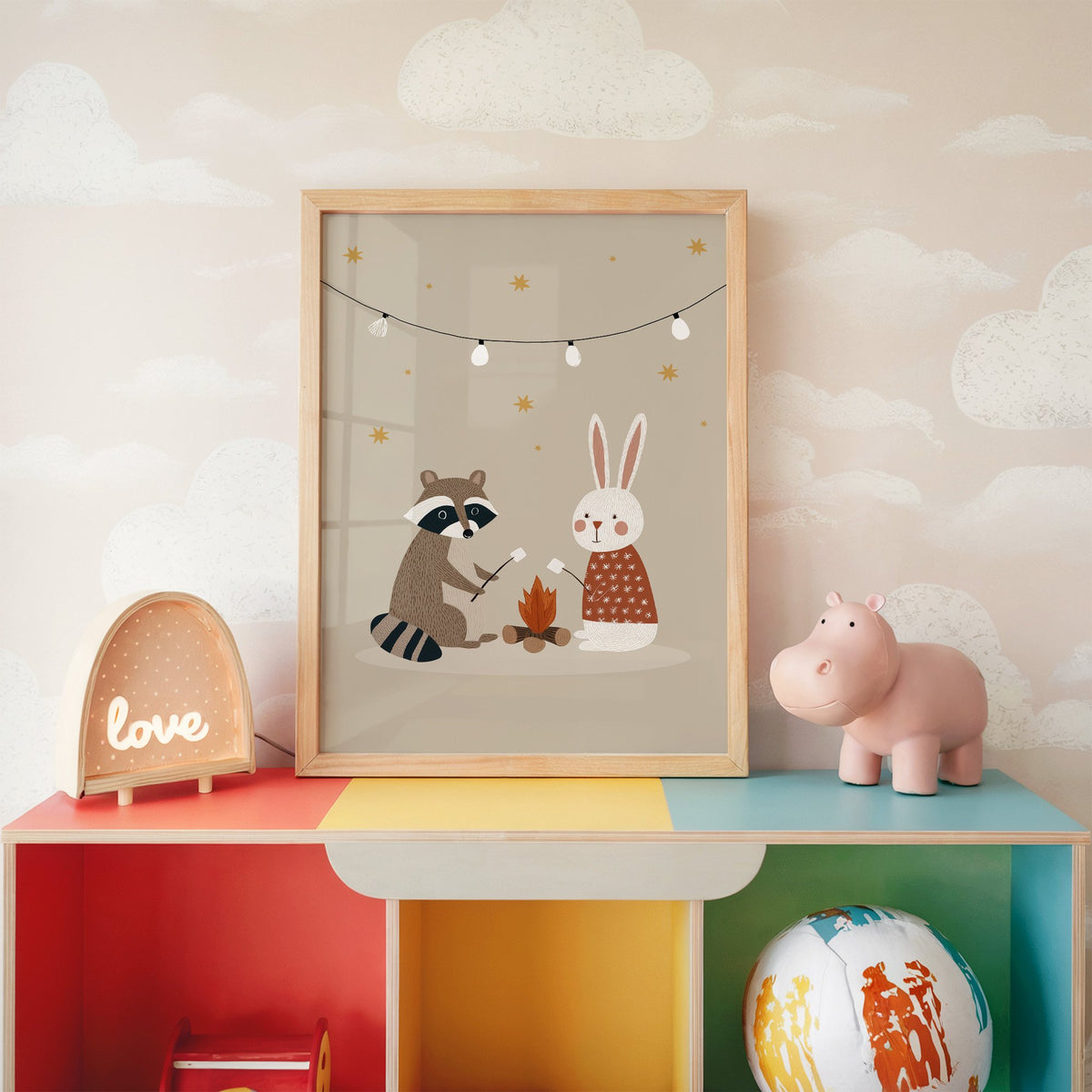 Raccoon and bunny campfire nursery print in an oak frame on a rainbow toy shelf against cloud wallpaper