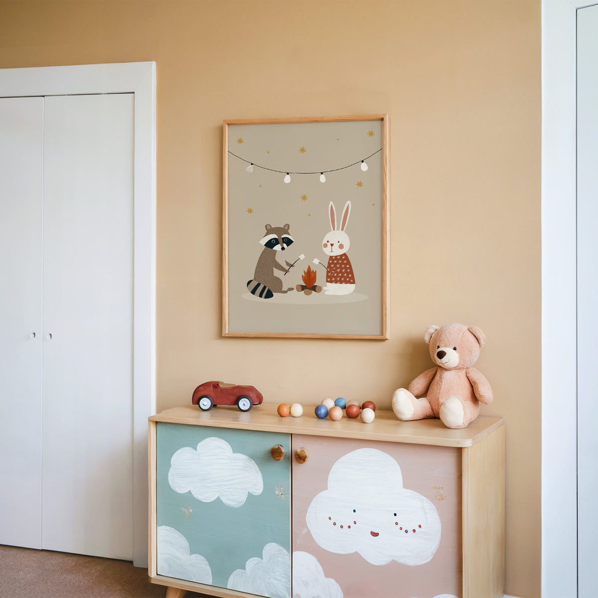 Raccoon and bunny campfire nursery print in an oak frame on a peach wall above a cloud-painted cabinet