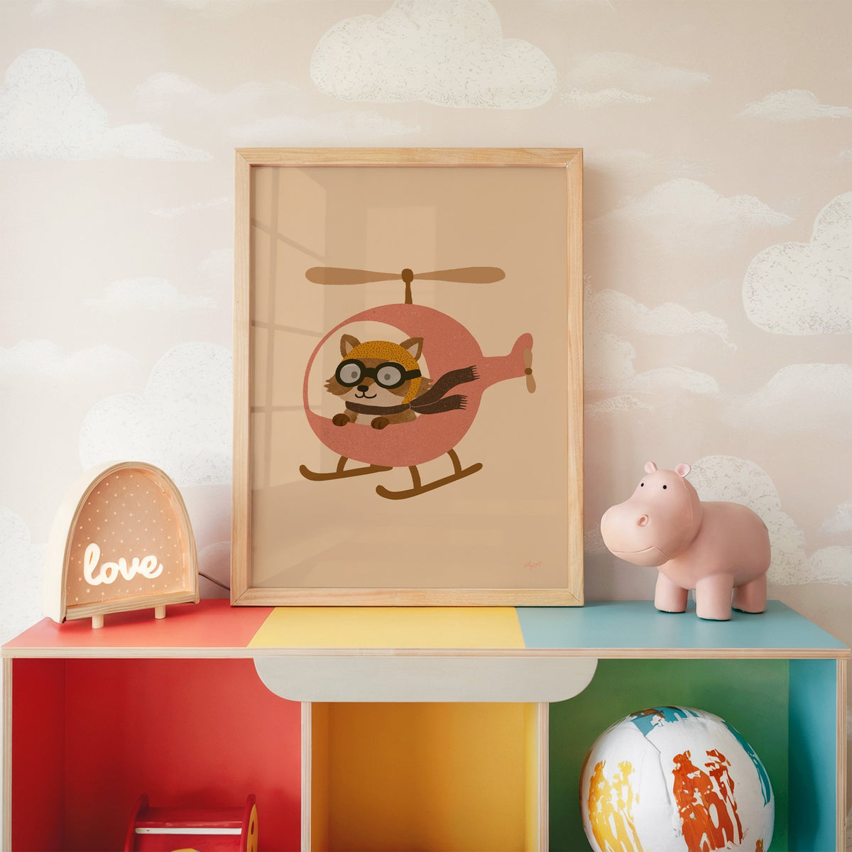 Raccoon pilot in pink helicopter nursery print in an oak frame on a rainbow toy shelf against cloud wallpaper