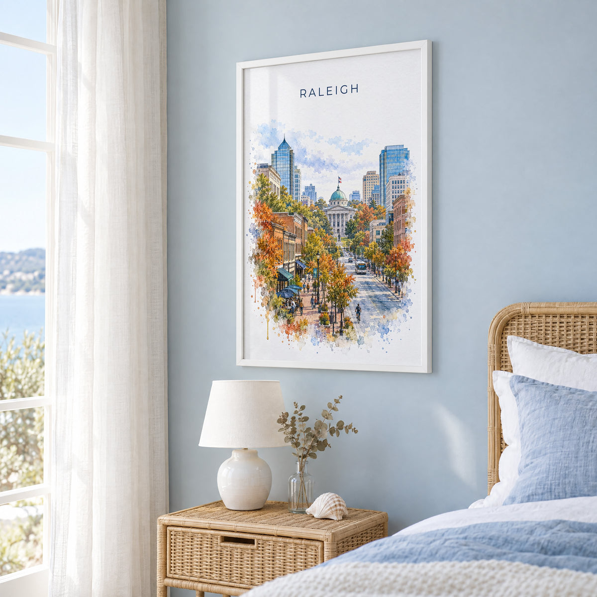 Raleigh watercolor print in a white frame on a pale blue wall beside a rattan bed in a coastal bedroom