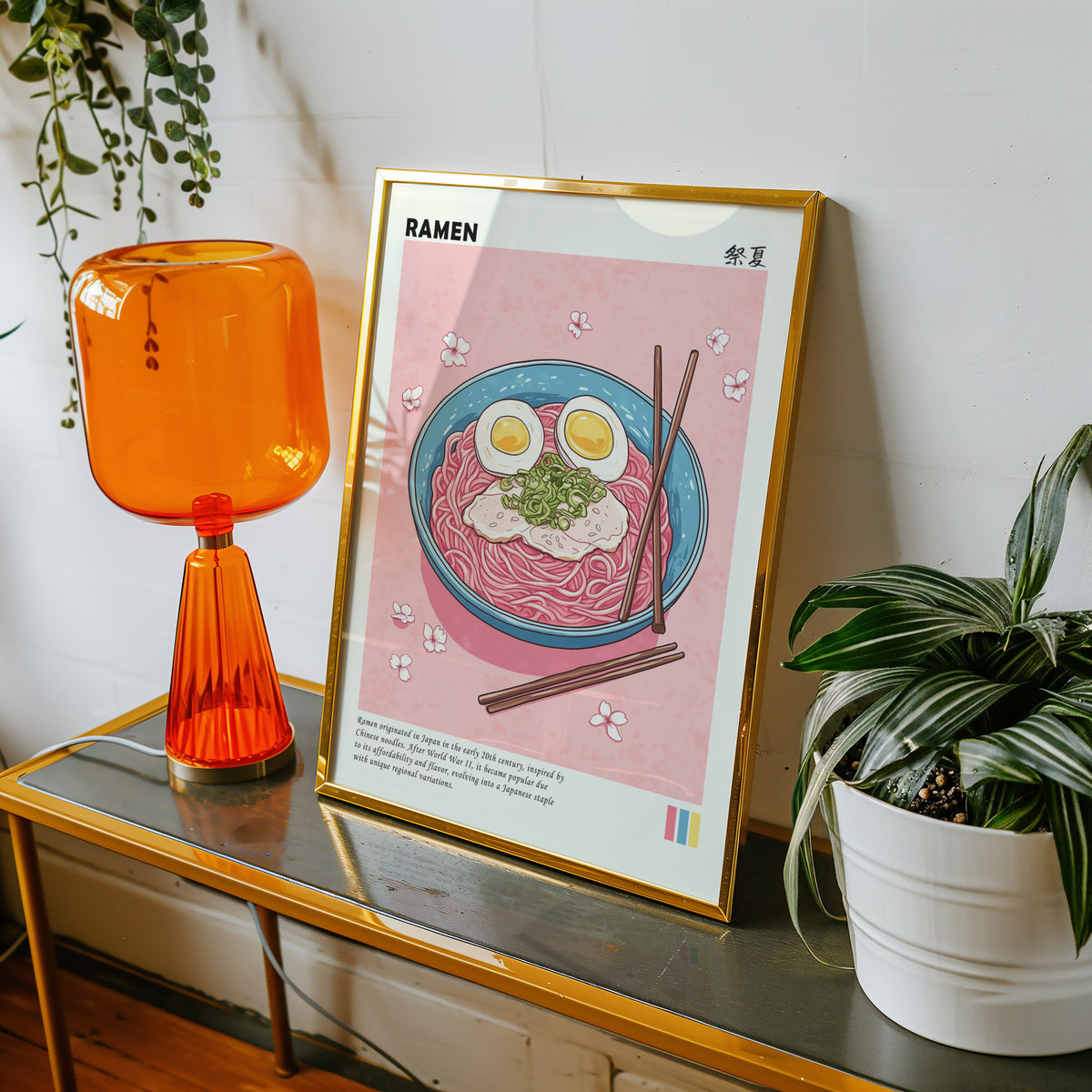 Turquoise ramen bowl with pink noodles print shown in a gold frame on a console beside an orange glass lamp