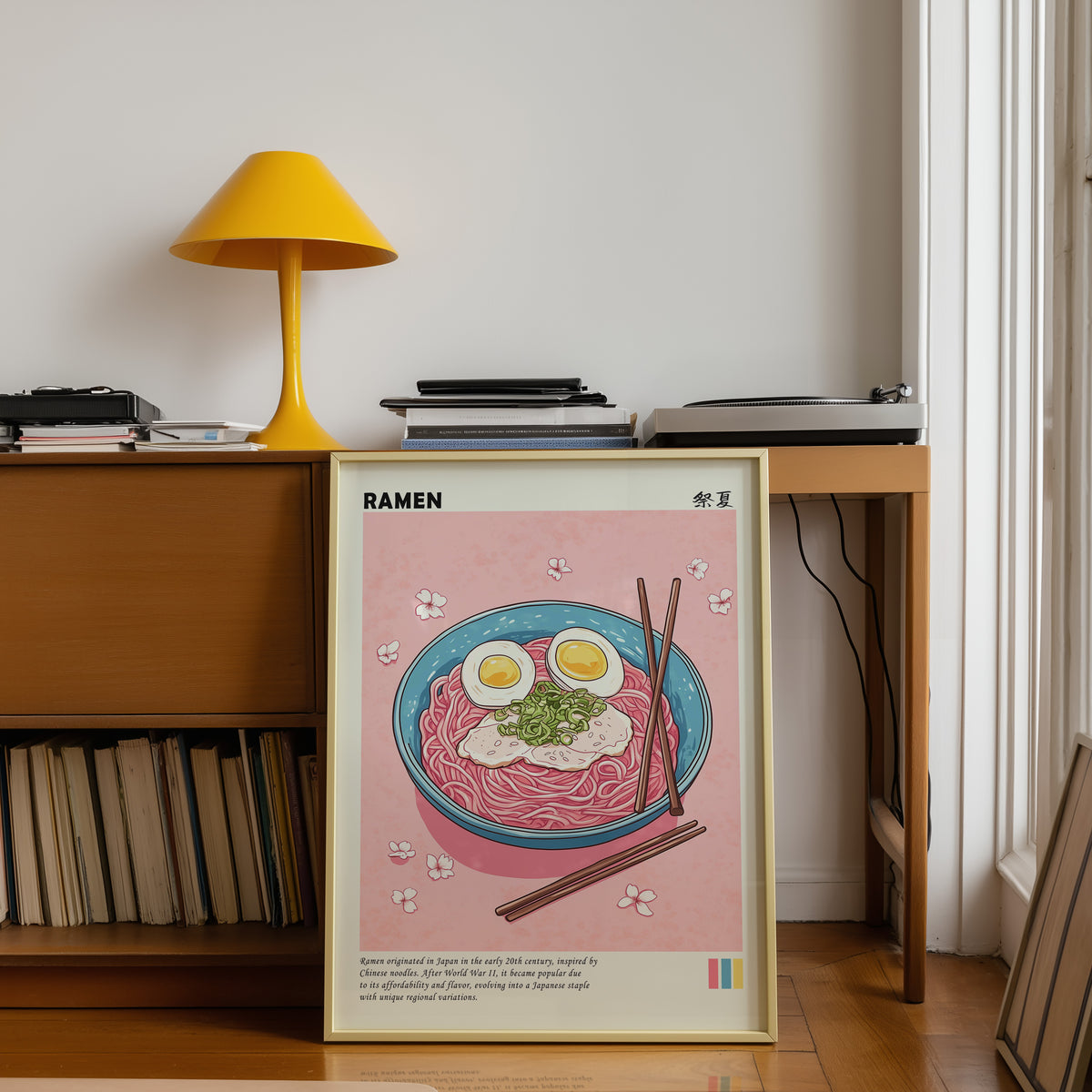 Turquoise ramen bowl with pink noodles print shown in a gold frame below a teak desk with a yellow lamp
