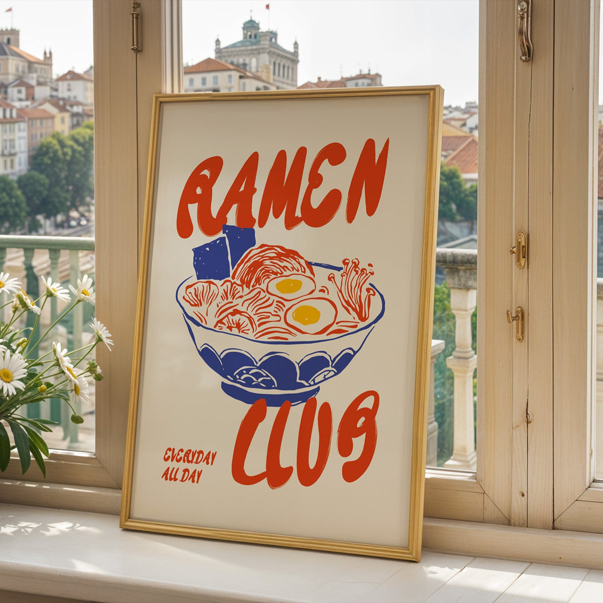 Ramen Club print in a gold frame on a window sill above a European hillside town