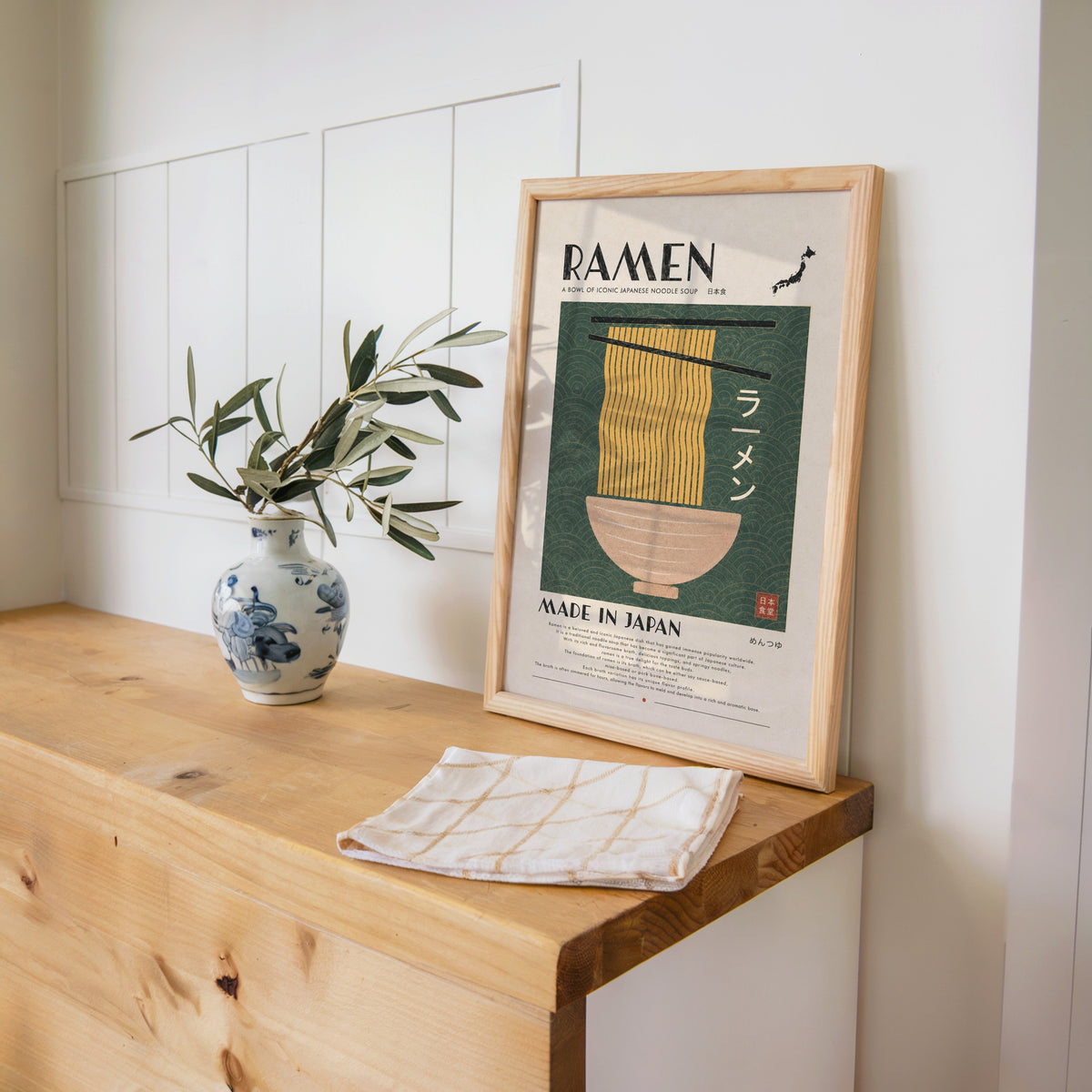 ramen noodles on green wave panel print in a pine frame on a wooden counter beside a blue and white porcelain vase
