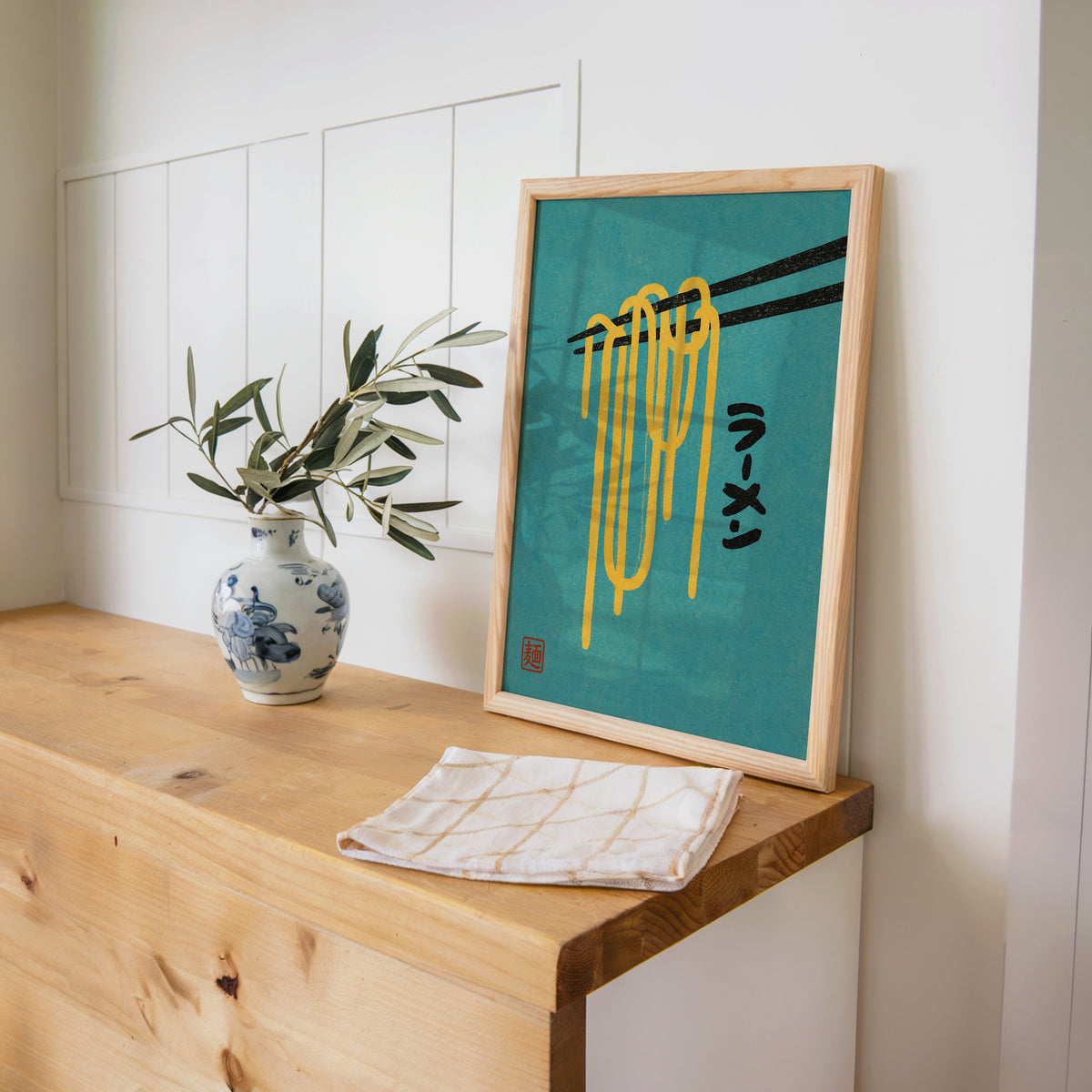 ramen noodles and chopsticks on teal print in a pine frame on a wooden counter beside a blue and white porcelain vase