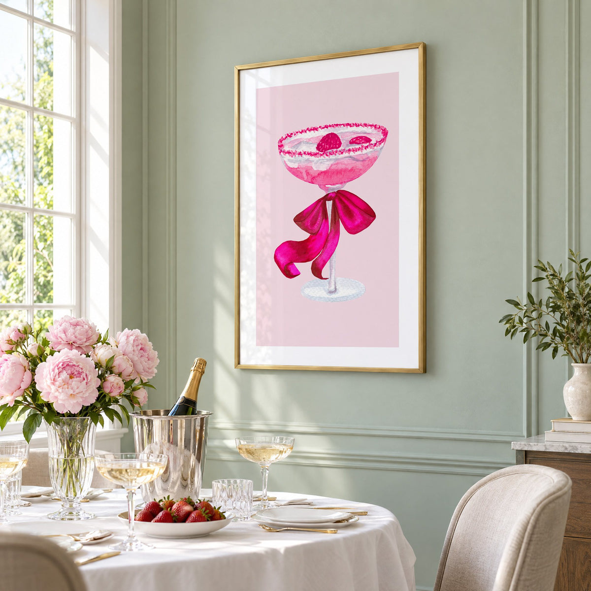 raspberry coupe magenta bow watercolor print in a brass frame on a sage wall above a brunch table with champagne and peonies, seen at an angle