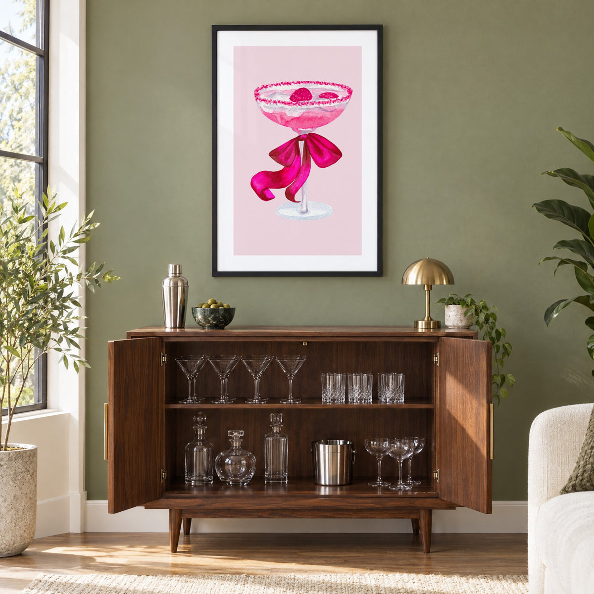 raspberry coupe magenta bow watercolor print in a black frame on an olive green wall above a walnut bar cabinet with glassware