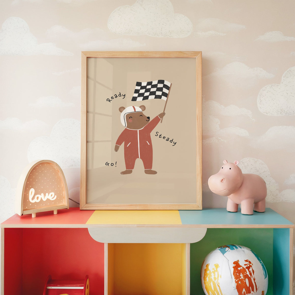 Ready steady go racing bear nursery print in an oak frame on a rainbow toy shelf against cloud wallpaper