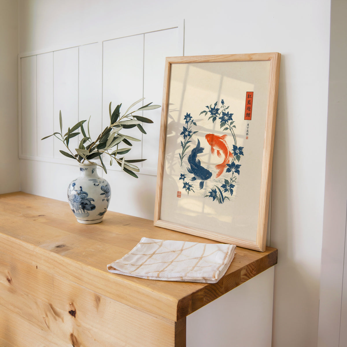 red and blue koi with blue flowers print in a pine frame on a wooden counter beside a blue and white porcelain vase
