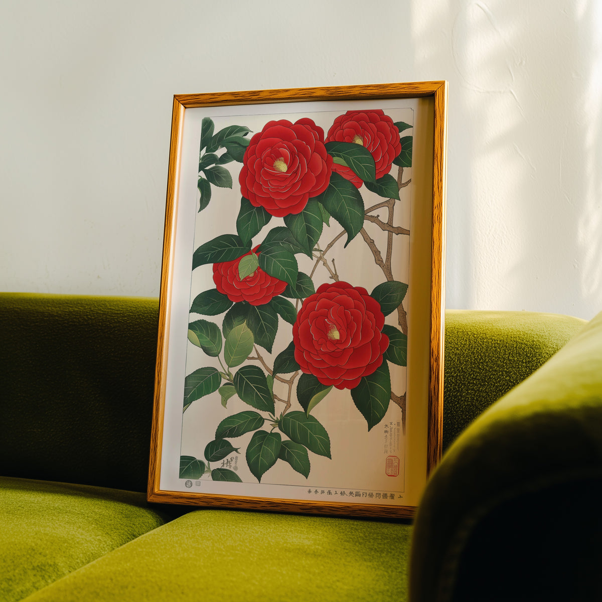 Red camellia woodblock-style print in an oak frame propped on a green velvet sofa