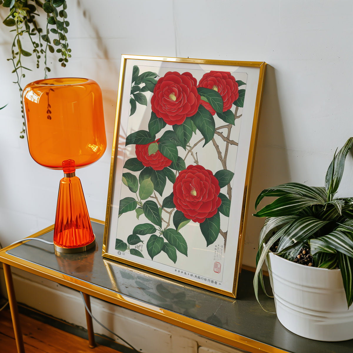 Red camellia woodblock-style print shown in a gold frame on a console beside an orange glass lamp