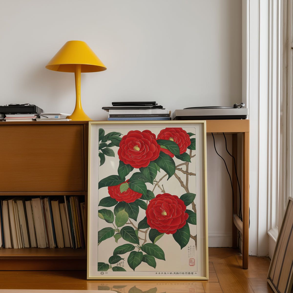 Red camellia woodblock-style print shown in a gold frame below a teak desk with a yellow lamp