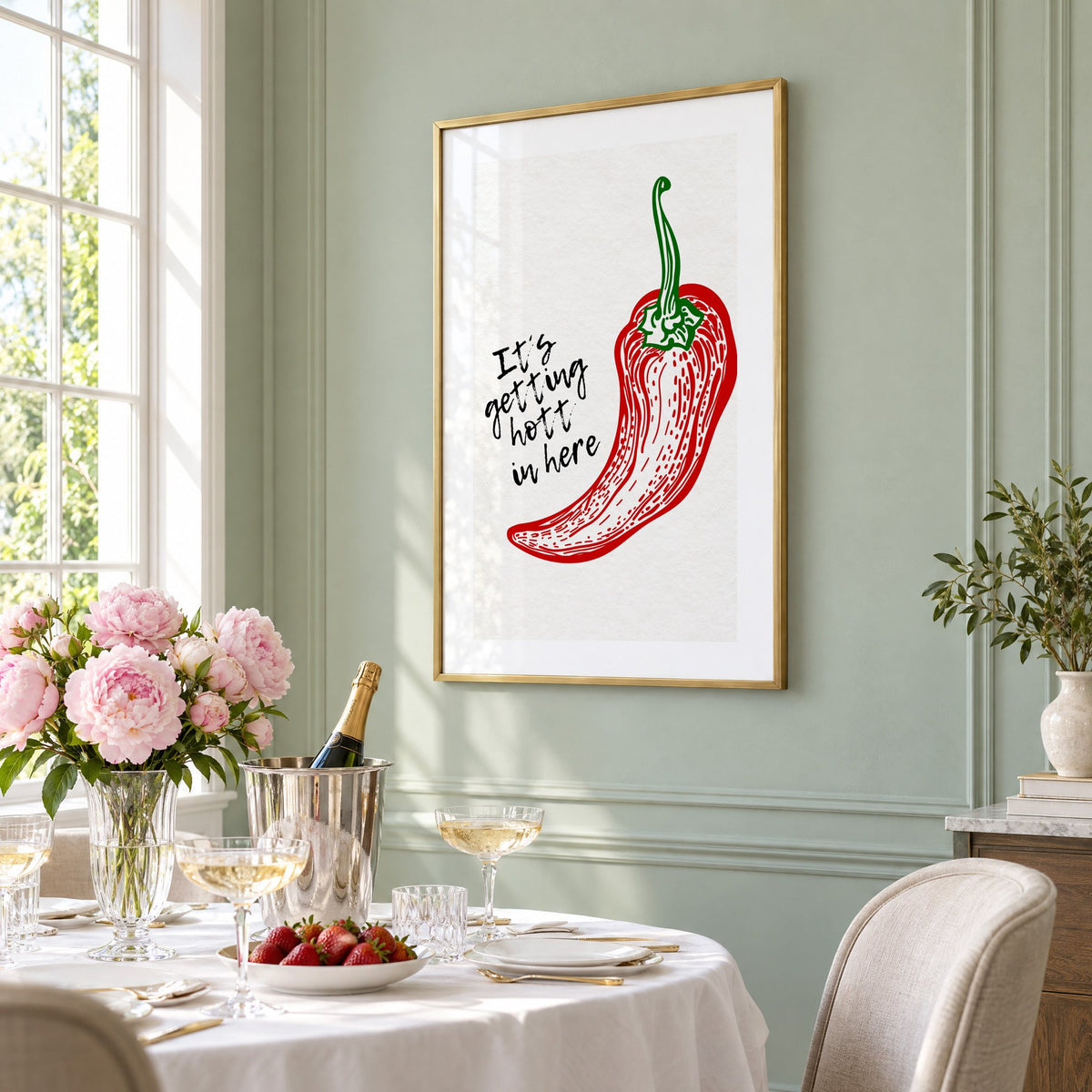 It's getting hott red chili engraving print in a brass frame on a sage wall above a brunch table with champagne and peonies, seen at an angle